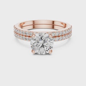 Puede incluir: Juego de anillos de compromiso de oro rosa con una gran piedra central de diamante de talla redonda. La banda presenta una doble fila de diamantes más pequeños y brillantes. El anillo está sobre un fondo blanco.
