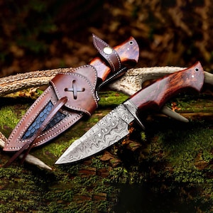 May include: A handcrafted hunting knife with a patterned steel blade and a wooden handle, resting beside its leather sheath. The sheath features a cross design and a decorative pattern. The knife and sheath are displayed on a mossy log.