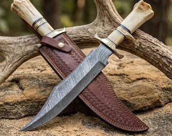 May include: Two hunting knives with Damascus steel blades and bone handles. The knives are in brown leather sheaths with decorative stitching. The handles have brass accents. The blades are crossed, resting on a stone surface.