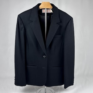 May include: A black blazer with a single-button closure, a notched lapel, and two front pockets. The blazer is hanging on a wooden hanger. The garment has a tailored fit and a classic design, suitable for formal or business attire.