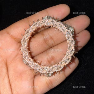 May include: A circular, rose gold-toned pendant with a crown of thorns design. The pendant is encrusted with small, sparkling crystals. The word "ICEFORGE" is printed in white in several places around the pendant.