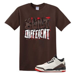 May include: A brown t-shirt with the words "BUILT DIFFERENT" in a graffiti-style design. A white, black, and brown sneaker with a red sole is positioned below the shirt.