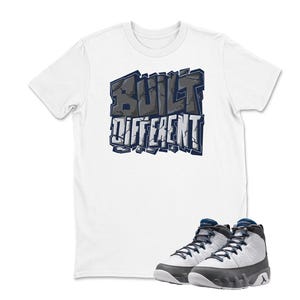 May include: White t-shirt with the text "BUILT DIFFERENT" in a blue and gray graffiti style. A pair of white and gray high-top sneakers with blue accents are in front of the shirt.
