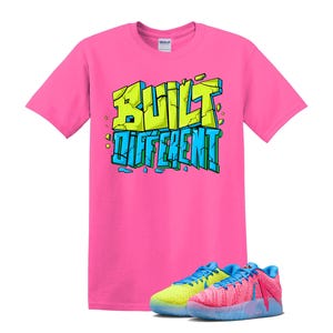 May include: A bright pink t-shirt with the words "BUILT DIFFERENT" in a graffiti-style design. Below the shirt are two pairs of sneakers, one pair is yellow and blue, and the other is pink and blue.