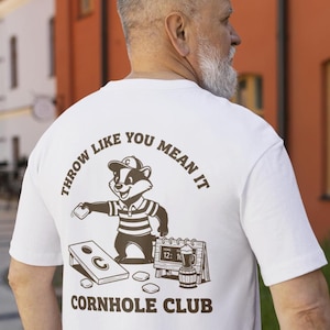 May include: White t-shirt with a brown graphic of a badger playing cornhole, the text "THROW LIKE YOU MEAN IT" and "CORNHOLE CLUB". The badger wears a hat and striped shirt. A cornhole board and beer barrel are also shown.