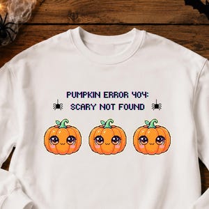 Op de afbeelding: Een wit sweatshirt met lange mouwen voorzien van een pixel art afbeelding van drie oranje pompoenen met grote ogen en de tekst PUMPKIN ERROR 404: SCARY NOT FOUND, geflankeerd door twee kleine zwarte spinnen aan weerszijden van de tekst.