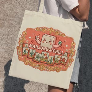 Puede incluir: Bolso tote de color crema con una ilustración colorida. El diseño incluye un personaje de ficha de mahjong sonriente y el texto "MAHJONG & CHILL" en un marco decorativo con azulejos de paisaje desértico. El bolso lo sostiene una persona.