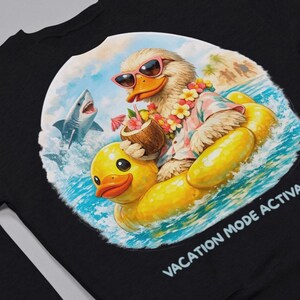 Op de afbeelding: Zwart sweatshirt met een kleurrijke afbeelding van een eend met zonnebril en een Hawaiiaans shirt, drijvend op een gele opblaasbare eend. Een haai is zichtbaar. De tekst luidt "VACATION MODE ACTIVATED".