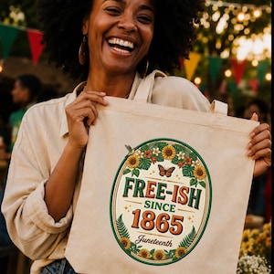 Op de afbeelding: Een beige draagtas met de tekst "FREE-ISH SINCE 1865 Juneteenth" in een cirkelvormig ontwerp met zonnebloemen, vlinders en varens. De tas wordt vastgehouden door een persoon die een beige shirt en een blauwe spijkerbroek draagt.