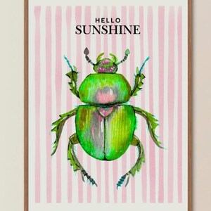 Op de afbeelding: Een ingelijste kunstprint met een levendige groene kever gecentreerd op een achtergrond van verticale lichtroze strepen. De tekst HELLO SUNSHINE staat in zwarte hoofdletters boven de kever gedrukt. Het kunstwerk zit in een dunne houten lijst.