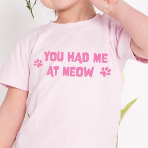 May include: A light pink t-shirt with the phrase "YOU HAD ME AT MEOW" in bright pink letters. The shirt also features pink paw prints on either side of the text, creating a playful design. The shirt is made of a soft material.