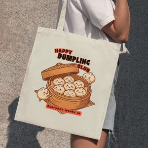 Op de afbeelding: Crèmekleurige draagtas met een cartoonillustratie van dumplings in een bamboestomer. De tekst "HAPPY DUMPLING CLUB" en "Everyone wants in" zijn ook aanwezig. De tas wordt door een persoon vastgehouden.