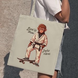 Op de afbeelding: Een beige tote bag met een grafische illustratie van een aap in een wit judopak met een zwarte band, staand op een skateboard. De tekst "Born to throw" en "Judo vibes" is in zwarte schrijfletters naast de aap op de tas gedrukt.