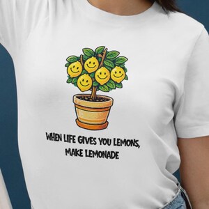 Op de afbeelding: Wit T-shirt met een cartoon citroenboom ontwerp in een terracotta pot. De citroenen hebben lachende gezichten. De tekst "WHEN LIFE GIVES YOU LEMONS, MAKE LEMONADE" staat eronder. Het shirt is gemaakt van zacht materiaal.
