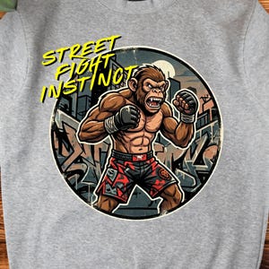 Op de afbeelding: Een grijs gemêleerd sweatshirt met ronde hals en een ronde afbeelding van een gespierde chimpansee in vechthouding. De aap draagt een zwart-rode broek met patroon en zwarte handschoenen. De gele tekst luidt STREET FIGHT INSTINCT.