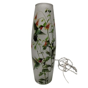 May include: A tall, off-white lamp with a nature-themed design. The lamp features illustrations of hummingbirds, red flowers, and green foliage. The lamp has a cylindrical shape and a textured surface. A white electrical cord extends from the base.