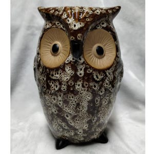 May include: A ceramic owl figurine with a speckled brown and white glaze. The owl has large, tan-colored eyes with black pupils and a black beak. The figurine is standing upright on a white surface.