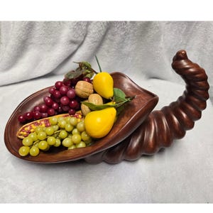 May include: A brown, horn-shaped cornucopia bowl filled with artificial fruit, including clusters of red and green grapes, two bright yellow pears, and several brown walnuts, all arranged together within the curved, textured vessel.