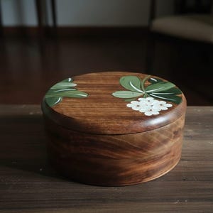 May include: A round wooden box with a lid. The box is stained a warm brown color. The lid is decorated with green leaves and white flowers. The box is sitting on a wooden surface.
