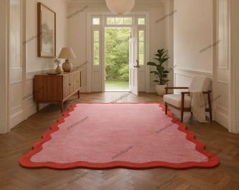 May include: A large, rectangular rug with a wavy, scalloped edge. The rug is primarily a soft pink color with a vibrant red border. The rug is placed in a hallway with a wooden floor and a white wall.