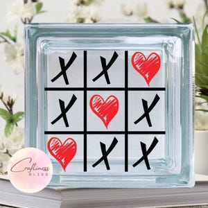 May include: A glass block with a tic-tac-toe game design. The game board is filled with red hearts and black X's. The hearts are hand-drawn and the X's are bold and black.