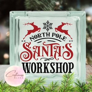 May include: A glass block with a red and black design that says "North Pole Santa's Workshop". Two red reindeer silhouettes are on either side of the text. A snowflake is above the text.