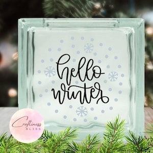 May include: A glass block with a white background and blue snowflakes and dots. The text "hello winter" is written in black cursive lettering.