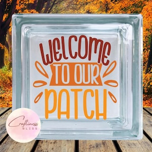 May include: A glass block with the words "Welcome to our Patch" in orange and brown lettering. The block is sitting on a wooden surface with a blurred background of fall foliage.