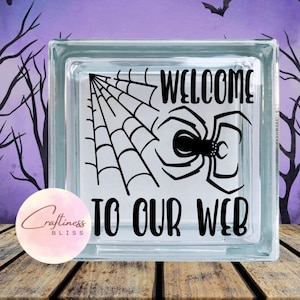 May include: A clear glass block with a black spider web and spider design. The text "WELCOME TO OUR WEB" is displayed in black. The background features a Halloween theme with bats and bare trees.
