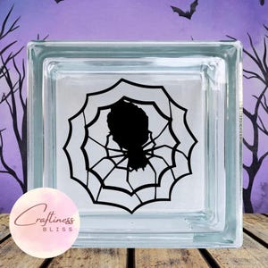 May include: A clear glass block with a black silhouette of a spider in a web. The glass block is on a wooden surface.