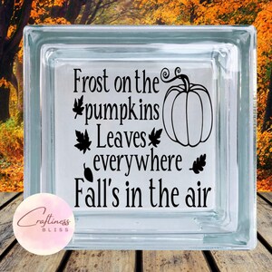 Op de afbeelding: Een helder glazen blok met de tekst "Frost on the pumpkins, Leaves everywhere, Fall's in the air" en een pompoenafbeelding in zwart. Geplaatst op een houten oppervlak met een vage herfstachtergrond.