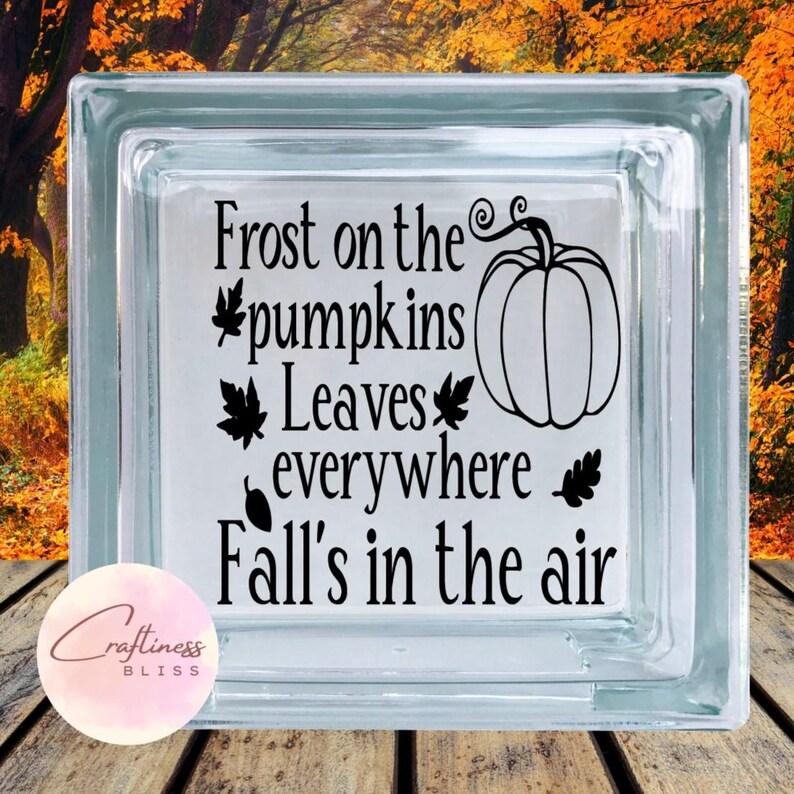Op de afbeelding: Een helder glazen blok met de tekst "Frost on the pumpkins, Leaves everywhere, Fall's in the air" en een pompoenafbeelding in zwart. Geplaatst op een houten oppervlak met een vage herfstachtergrond.