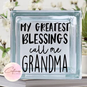 May include: A clear glass block with the text "MY GREATEST BLESSINGS call me GRANDMA" in black lettering. The block is sitting on a stack of books. A Craftiness Bliss logo is in the lower left corner.