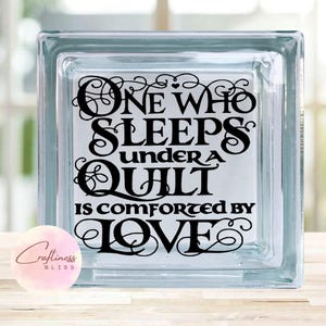 May include: A glass block with a black vinyl decal that reads "One who sleeps under a quilt is comforted by love".
