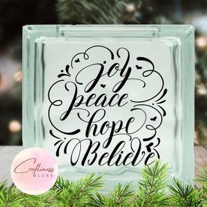 May include: A clear glass block with the words "Joy, Peace, Hope, Believe" written in black cursive script. The words are surrounded by decorative swirls and hearts.
