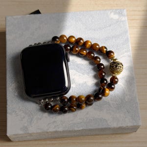 May include: A black smartwatch with a beaded bracelet. The bracelet is made of brown and gold tiger eye beads and features a decorative gold-coloured charm. The watch and bracelet are displayed on a light grey surface.