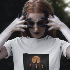 Pode incluir: Camiseta branca com uma ilustração escura de três bruxas em vestidos e chapéus pretos em frente a uma lua cheia. Abaixo das bruxas estão vários esqueletos. A imagem tem um tema assustador de Halloween.