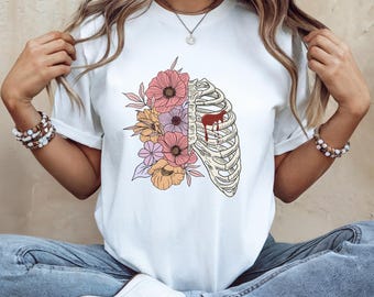 May include: White t-shirt featuring a graphic design of a rib cage with colorful flowers on the left side and a red heart-shaped element. The flowers are in shades of pink, orange, and purple.
