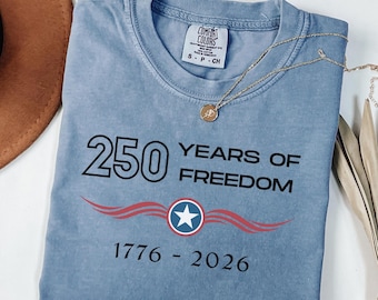 May include: A blue Comfort Colors t-shirt with the text "250 YEARS OF FREEDOM 1776-2026" in black. The design includes a red and blue graphic with a star. A gold necklace is draped across the shirt.