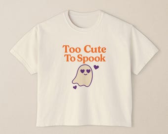 Puede incluir: Camiseta color crema con el texto "Too Cute To Spook" en naranja y un simpático dibujo de un fantasma. El fantasma tiene ojos de corazón morados y está rodeado de corazones morados. Una camiseta divertida con temática de Halloween.