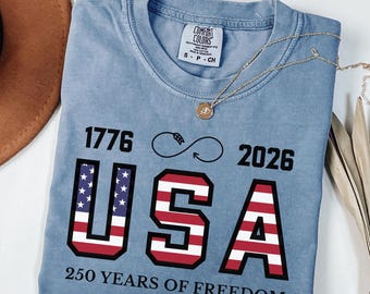May include: Blue jean colored t-shirt with the text "1776 USA 2026 250 YEARS OF FREEDOM" in red, white, and blue. The shirt has a crew neck and short sleeves. A gold necklace with a pendant is draped across the shirt.