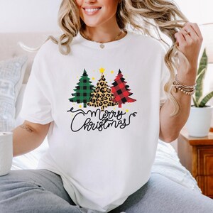 Może przedstawiać: Biała koszulka z trzema choinkami w zieloną kratę, lamparcie cętki i czerwono-czarną kratę. Pod choinkami napis "Merry Christmas" w ozdobnej czcionce.