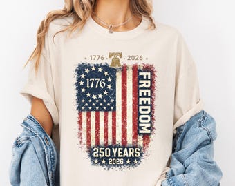 May include: Cream-colored t-shirt featuring a distressed American flag design. The flag includes the text "1776" and "FREEDOM", with "250 YEARS 2026" below. The shirt has a vintage, patriotic theme.