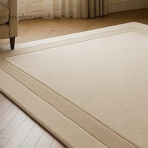 May include: A rectangular beige rug with a bordered design, placed on a wooden floor. The rug has a lighter beige center and a darker beige border. A beige armchair is partially visible in the upper left corner.