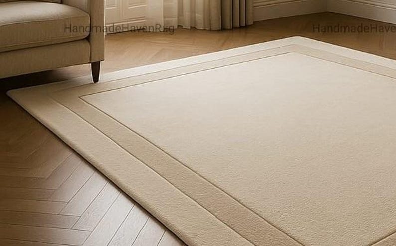 May include: A rectangular beige rug with a bordered design, placed on a wooden floor. The rug has a lighter beige center and a darker beige border. A beige armchair is partially visible in the upper left corner.