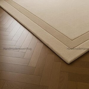 May include: A beige rectangular rug with a geometric border is placed on a wooden parquet floor. The rug's texture is visible, and the corner is in focus. The words "HandmadeHavenRug" are printed on the rug.