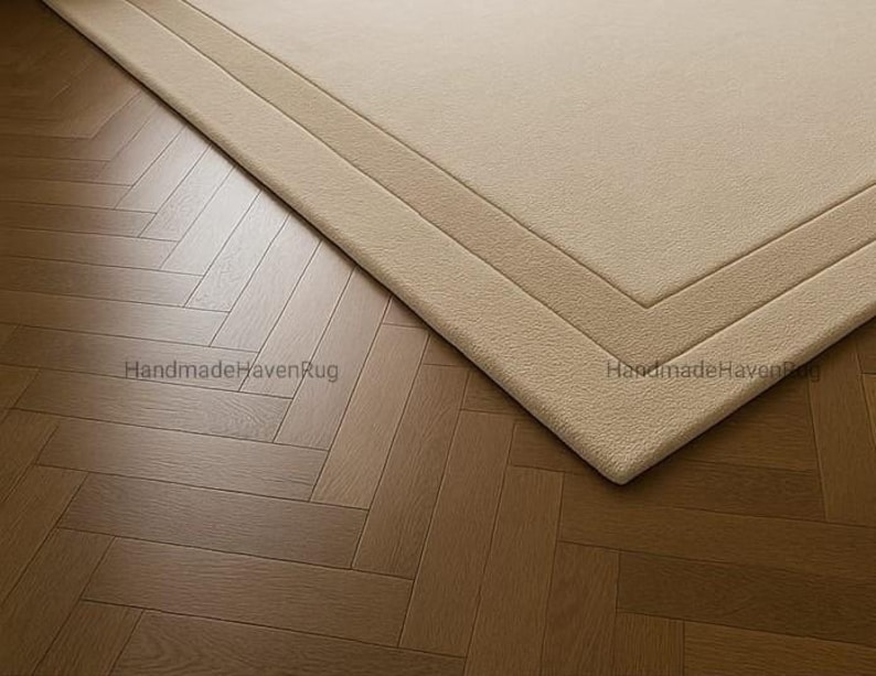 May include: A beige rectangular rug with a geometric border is placed on a wooden parquet floor. The rug's texture is visible, and the corner is in focus. The words "HandmadeHavenRug" are printed on the rug.