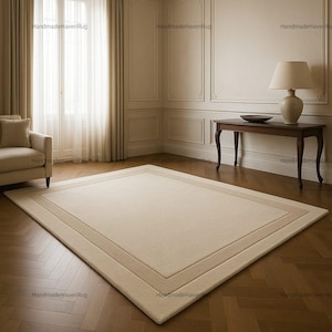 May include: A rectangular beige rug with a lighter beige border on a parquet floor. The room features a window with curtains, a side table with a lamp, and decorative wall panels. The overall style is classic and elegant.