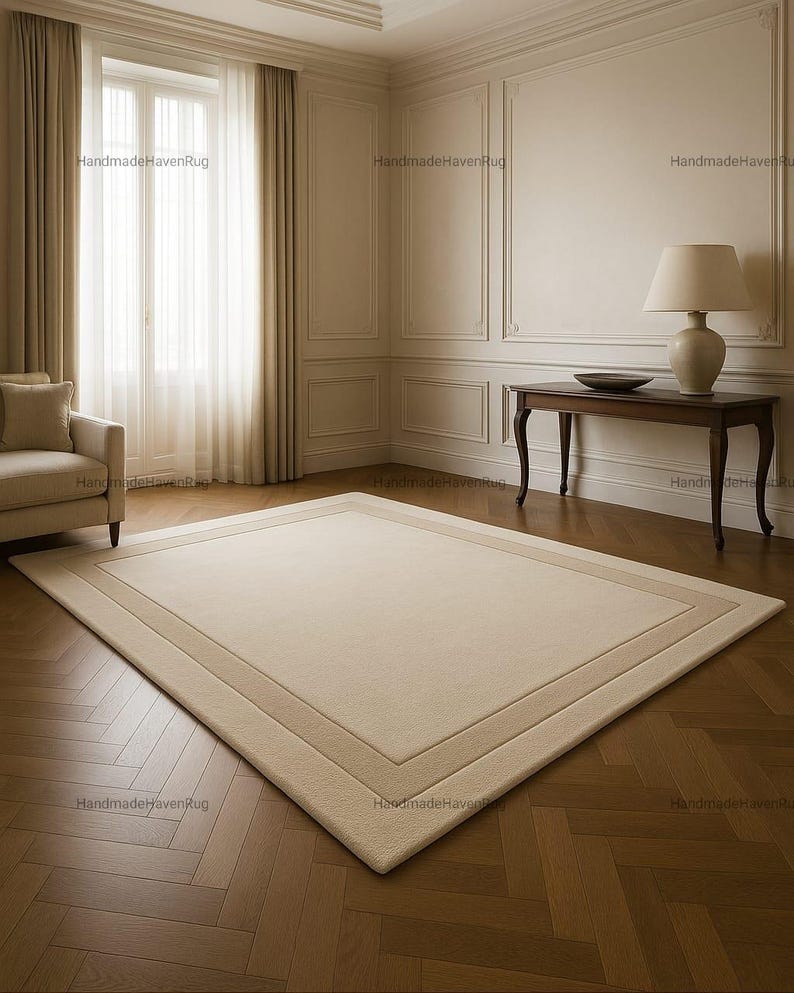 May include: A rectangular beige rug with a lighter beige border sits on a hardwood floor. The rug is in a room with a window, curtains, and a side table with a lamp. The room has a classic, elegant style.