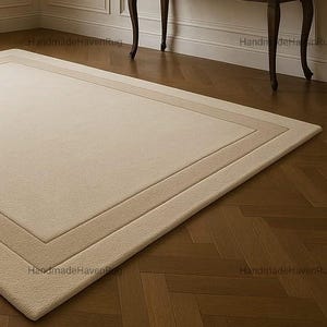 May include: A rectangular beige rug with a bordered design, placed on a herringbone pattern hardwood floor. The rug has a soft, plush texture. A dark brown table with curved legs and a decorative vase are in the background.